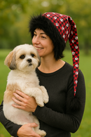 Woman in a black sweater holding a small fluffy dog while wearing a handmade red plaid hat with white paw prints, black fuzzy trim, and a long drooping tail with a black pom-pom.