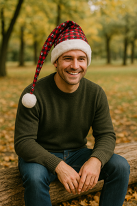 A smiling man wearing a red plaid Santa-style hat with white paw prints, white fur trim, and a fluffy pom-pom sits on a log in a park surrounded by autumn leaves.