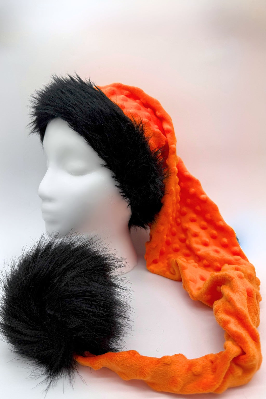 A bright orange textured winter (Ssanta-style) hat with a long tail, trimmed in fluffy black faux fur around the brim and finished with a large black faux fur pom-pom, displayed on a white mannequin head against a light background.