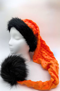 A bright orange textured winter (Ssanta-style) hat with a long tail, trimmed in fluffy black faux fur around the brim and finished with a large black faux fur pom-pom, displayed on a white mannequin head against a light background.