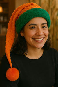 Smiling Latina woman wearing a handmade Orange Bumps holiday hat with green trim, orange bumpy fabric, and an orange pom-pom, sitting in a cozy coffee shop with coenstocks in the background.