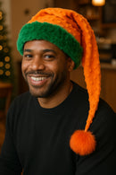 African American man wearing a handmade Orange Bumps holiday hat with green trim, orange bumpy fabric, and an orange pom-pom, sitting and smiling in a cozy coffee shop.

