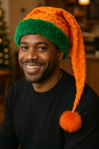 African American man wearing a handmade Orange Bumps holiday hat with green trim, orange bumpy fabric, and an orange pom-pom, sitting and smiling in a cozy coffee shop.
