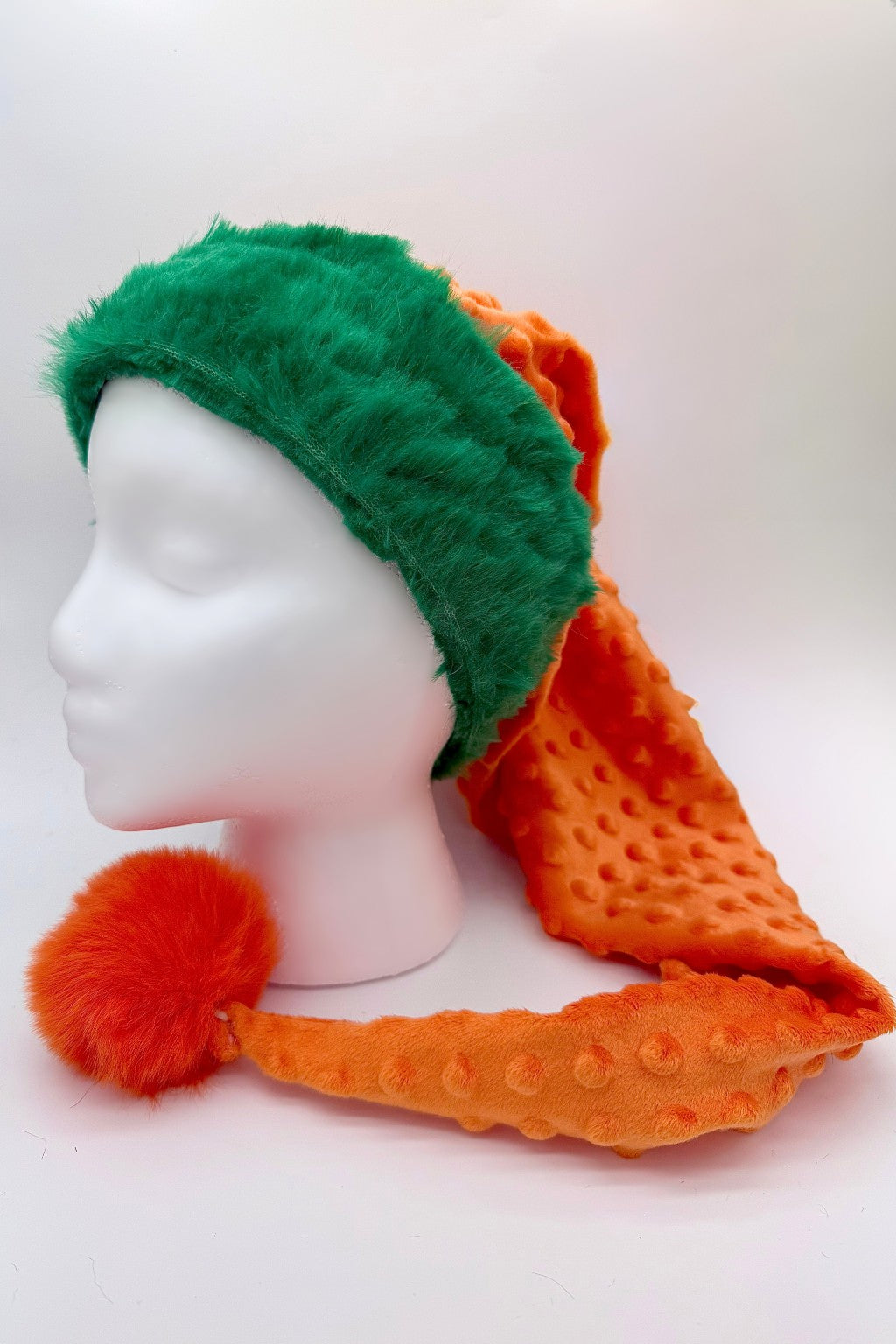 A mannequin head displays a whimsical (Ssanta-style) hat with a fuzzy green band and a long, textured orange tail made of dimpled fabric, ending in a fluffy orange pom-pom.