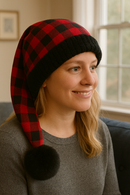 Blonde woman in a gray sweater wearing a red and black buffalo plaid hat with black faux fur trim and pom, sitting indoors by a window, mirrored view.