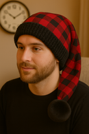Man with short light brown hair and a beard wearing a red and black buffalo plaid hat with black faux fur trim and pom, sitting indoors with a wall clock in the background.