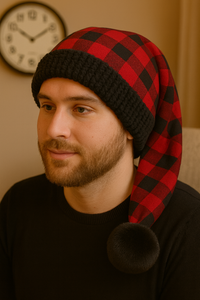 Man with short light brown hair and a beard wearing a red and black buffalo plaid hat with black faux fur trim and pom, sitting indoors with a wall clock in the background.