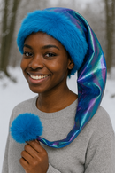 A young African woman smiles warmly outdoors in a snowy setting, wearing a shimmering iridescent hat with bright blue faux fur trim and a fluffy blue pom-pom. The hat is shorter in length, draping just past her shoulder, and she holds the pom-pom in her hand while dressed in a simple gray sweater.