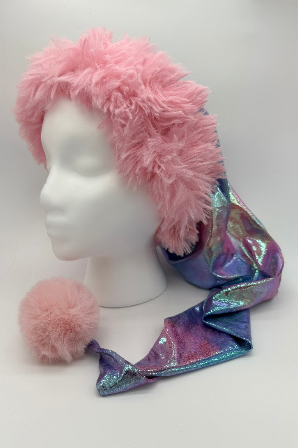 A fluffy pink faux fur hat with a long, iridescent pastel tie-dye tail ending in a matching pink pom-pom, displayed on a white mannequin head.