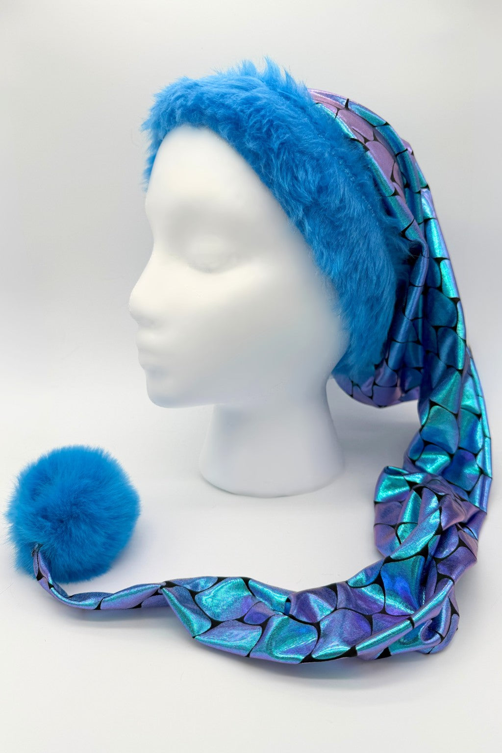 A mannequin head wearing a long, pointed hat with a wide band of bright blue faux fur. The main part of the hat is made of shiny, iridescent fabric in a purple and Teal mermaid-scale pattern, ending in a large fluffy blue pom-pom.