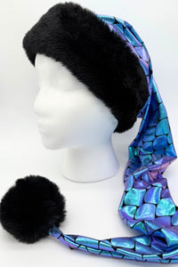 A mannequin head wearing a long, pointed hat with a wide black faux-fur brim and a shiny, iridescent fabric in blue and purple with a scale-like pattern. The hat extends down to the side, ending in a large black faux-fur pompom.
