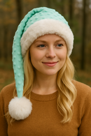 The image shows a young woman with long, wavy blonde hair wearing a light aqua Santa-style hat made of textured fabric with raised bumps. The hat is trimmed with white faux fur and finished with a large fluffy white pom-pom. She is dressed in a brown sweater and is outdoors, with a softly blurred background of trees and autumn foliage.