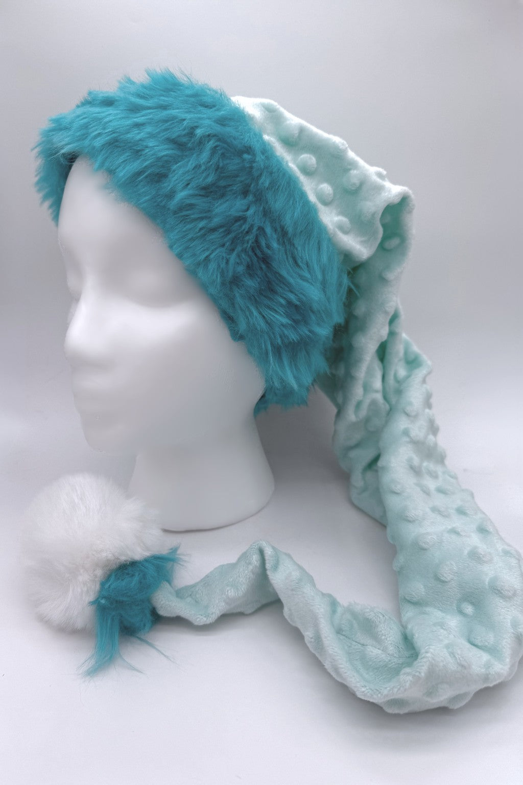 Light aqua blue textured fleece hat with a long tail, trimmed with bright Teal faux fur around the brim, and ending in a fluffy white pom-pom with Teal accents, displayed on a white mannequin head.