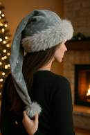 "A young woman with long brown hair wears a gray Velvet hat trimmed with fluffy light gray fur and decorated with jewels. She is shown from behind in a cozy indoor setting with a glowing fireplace and a blurred Christmas tree in the background, holding the hat’s long tail with a fur pom-pom."