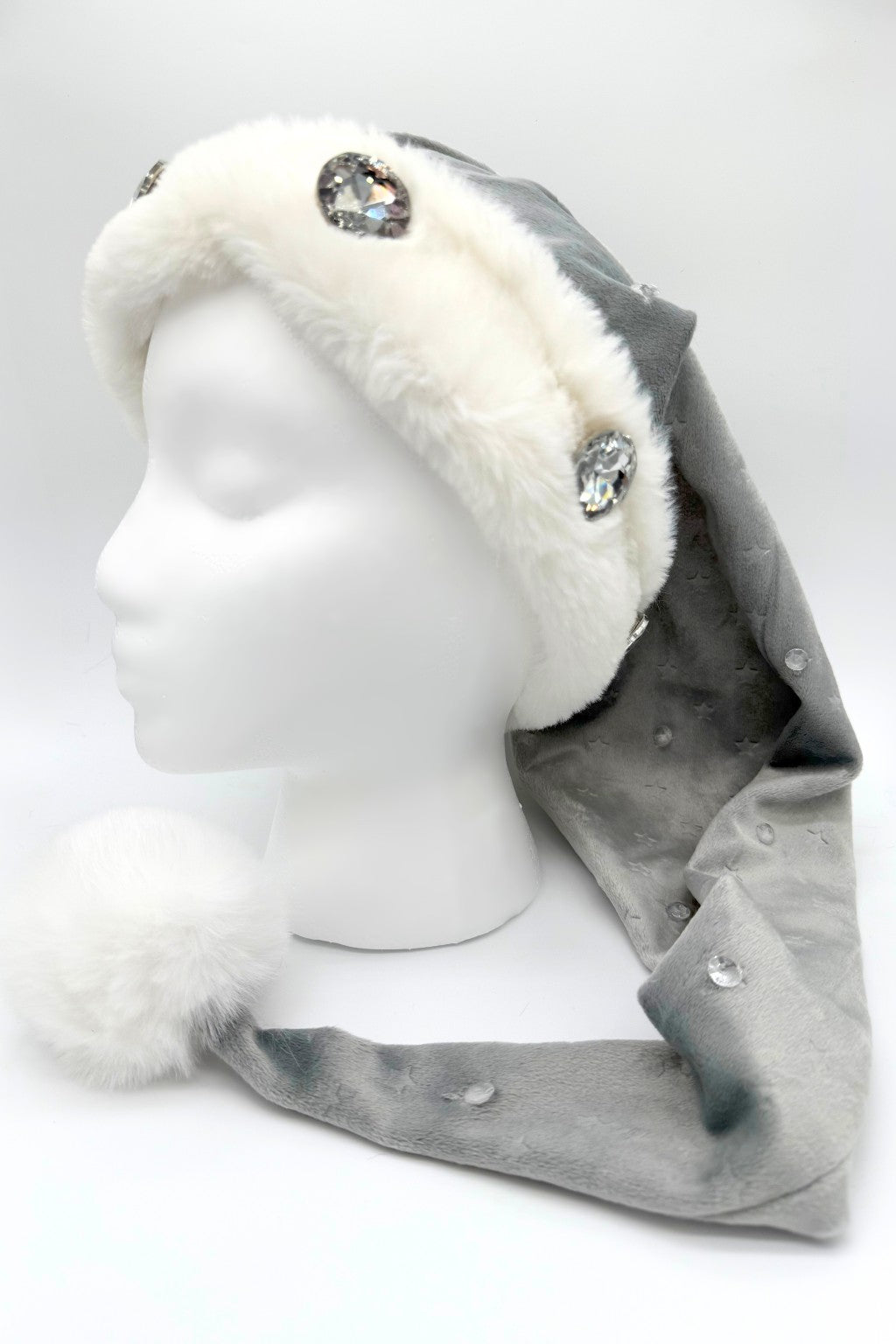 Gray Velvet Santa-style hat of silvery gray Velvet with embossed stars has a white faux fur trim and large rhinestone embellishments, and other more ssubtle jewel accents is finished with a fluffy white pom-pom at the tip, displayed on a white mannequin head.