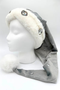Gray Velvet Santa-style hat of silvery gray Velvet with embossed stars has a white faux fur trim and large rhinestone embellishments, and other more ssubtle jewel accents is finished with a fluffy white pom-pom at the tip, displayed on a white mannequin head.