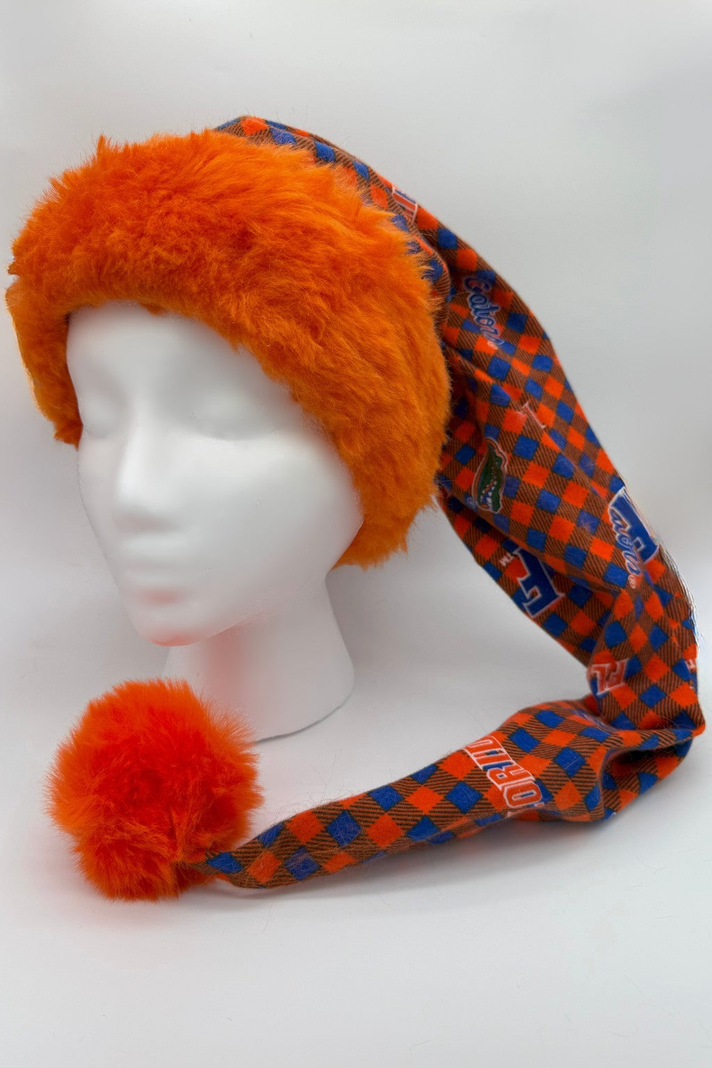 A mannequin head wearing a festive hat with bright orange faux fur trim and pom-pom. The long, pointed body of the hat is patterned in blue and orange checkered fabric with Florida Gators logos.