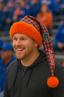 A smiling Florida Gators fan at a football game wearing an orange faux fur–trimmed Santa-style hat with a blue and orange checkered tail, the Gators logo, and “FLORIDA” lettering. The hat ends with a large orange pom-pom, and the fan is dressed in a dark zip-up hoodie, with blurred spectators in team colors in the background.