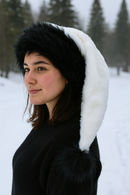 A young woman with fair skin and dark brown hair is standing outdoors in a snowy landscape, wearing a long white Velvet hat with a black faux fur trim and a black faux fur pom-pom at the end. She is dressed in a black sweater, and evergreen trees are visible in the background.