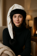 Woman wearing a white Velvet hat with black faux fur trim and black pom-pom, sitting indoors by a fireplace with a cozy blanket draped nearby.