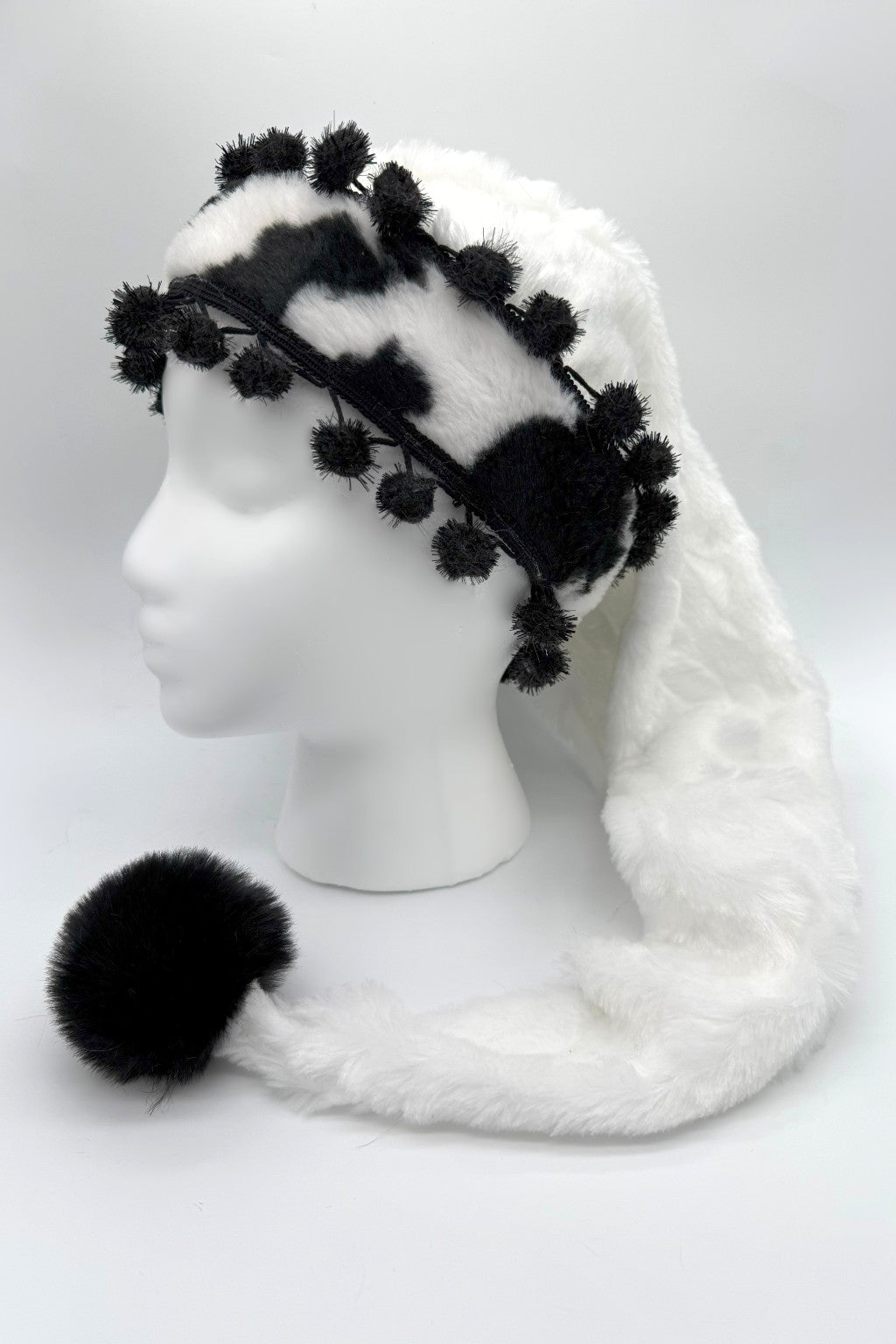 White faux fur hat with a long tail ending in a large black pom-pom, featuring a black-and-white cow print trim decorated with small black pom-poms, displayed on a white mannequin head.