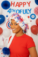 The image shows a young woman at a Fourth of July celebration, smiling while wearing a white faux fur hat adorned with red and blue tulle ribbons and star-shaped accents. The hat has a long tail ending in a fluffy blue pom-pom that drapes down one side. She is dressed in a plain white T-shirt, standing in front of a festive backdrop decorated with American flags, red, white, and blue balloons, and patriotic bunting.