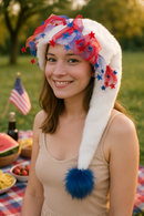 A young woman is sitting outdoors at a picnic on a checkered red, white, and blue blanket. She is smiling and wearing a festive white faux fur holiday hat decorated with red and blue star accents and sheer ribbon. The hat’s long tail extends down to rest just above her chest, ending with a fluffy blue pom-pom. In the background, picnic food such as watermelon, chips, and strawberries can be seen, along with an American flag, creating a cheerful holiday atmosphere.