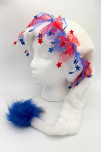 A white faux fur novelty hat displayed on a mannequin head. The hat features long ear flaps with a fluffy blue pom-pom at the end of one side. Around the crown, red and blue sheer ribbon is decorated with small red and blue star-shaped accents, giving the hat a festive, patriotic look.