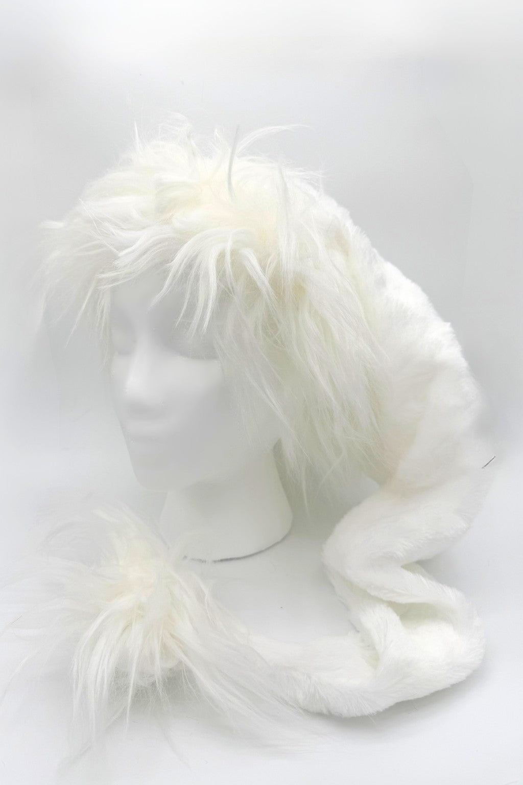 A fluffy white faux fur hat displayed on a white mannequin head. The hat has long, soft fur around the top and sides, with an extended scarf-like section that ends in a matching furry pom-pom.