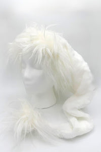 A fluffy white faux fur hat displayed on a white mannequin head. The hat has long, soft fur around the top and sides, with an extended scarf-like section that ends in a matching furry pom-pom.