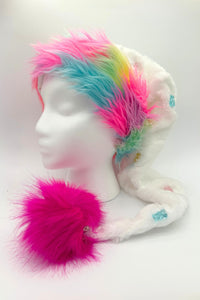 A white mannequin head wearing a whimsical hat with rainbow-colored faux fur trim in shades of pink, blue, green, and yellow. The hat is made of soft white fabric with small gummybear embellishments of multiple colors and ends in a long tail tipped with a large, fluffy, bright pink pom-pom.