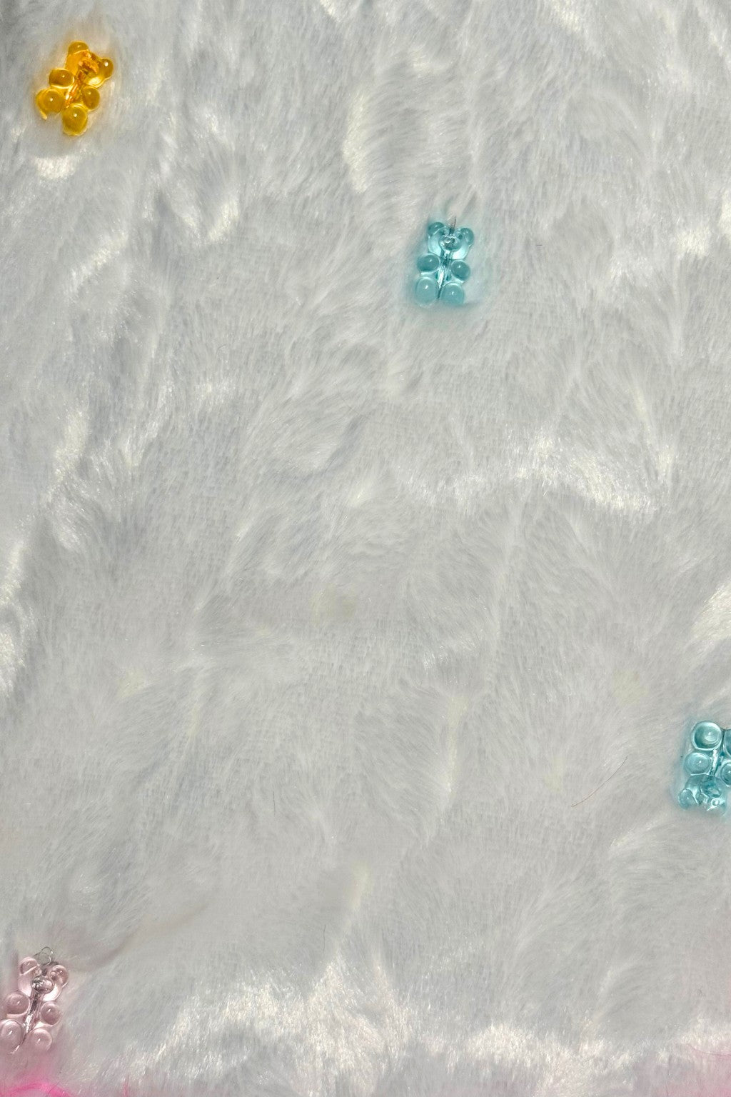 Colorful translucent teddy bear charms in yellow, blue, pink, and green scattered on a soft, white, furry fabric background.