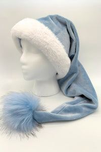 A light blue Velvet Santa-style hat with a fluffy white faux fur brim and a long tail ending in a large blue faux fur pom-pom tipped with gray. The hat is displayed on a white mannequin head against a plain background.