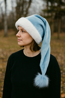 A young woman smiles outdoors, wearing a light blue Santa-style hat with white fur trim and a fluffy blue pom-pom, paired with a black sweater. The background is softly blurred with autumn trees and a path.