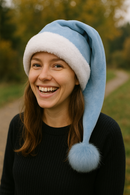 The image shows a young woman outdoors, smiling brightly while wearing a light blue Santa-style hat with fluffy white fur trim and a matching blue pom-pom. She has long brown hair, a black sweater, and the blurred background features autumn trees and a pathway.