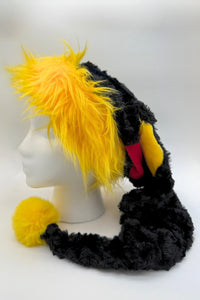 A black and yellow furry novelty (Santa-style) hat displayed on a white mannequin head. The hat features a bright yellow faux fur trim, long black plush sides that extend down, a red, blue and yellow diamond shaped insets, and a fluffy yellow pom-pom at the end of the hanging section.