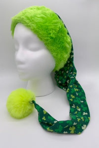 A festive green hat displayed on a white mannequin head. The hat has a bright neon-green furry brim and a long tail made from green fabric patterned with light and dark green shamrocks. The tail ends in a large fluffy neon-green pom-pom resting on the surface.
