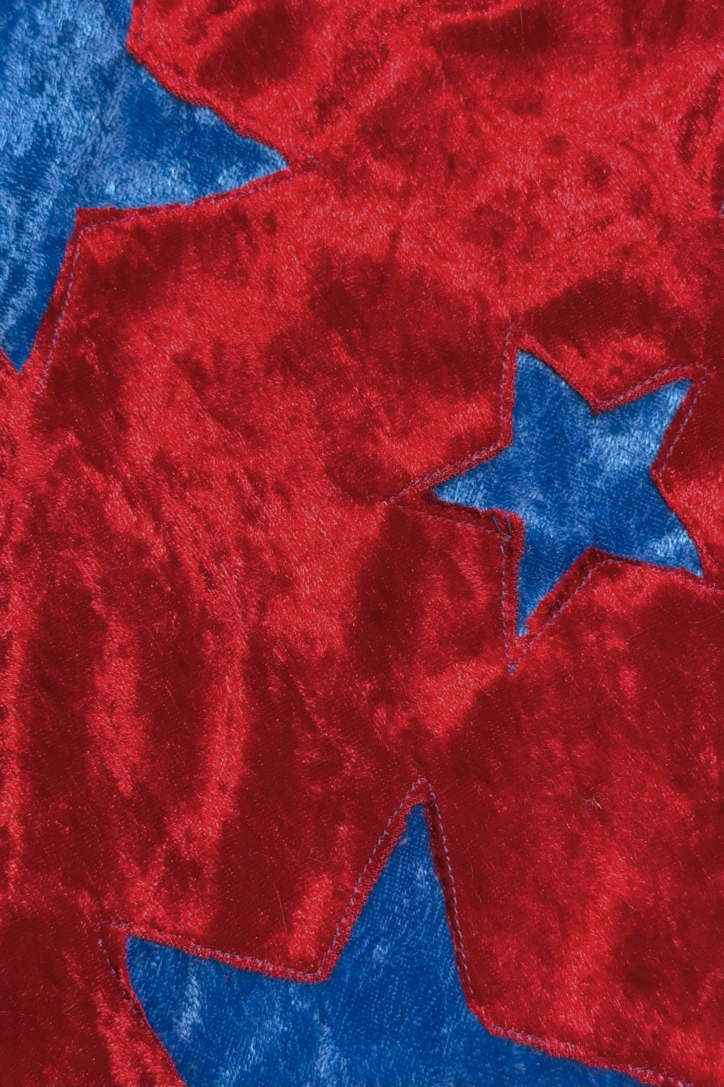 Close-up of a soft, shiny Velvet fabric with a red surface featuring cut-out star shapes, revealing a contrasting blue Velvet material underneath. The texture is smooth and reflective, giving a rich, glossy appearance.