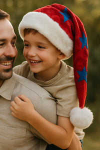 A smiling father carries his young son on his back outdoors, with the boy wearing a red Velvet Santa hat decorated with blue stars, white faux fur trim, and a fluffy pom-pom. Both share a joyful moment in the warm, golden sunlight with trees softly blurred in the background.