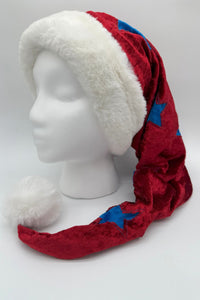A festive red Velvet Santa-style hat with blue star accents, trimmed with fluffy white faux fur, and finished with a large white pom-pom at the tip. The hat is displayed on a white mannequin head against a plain background.