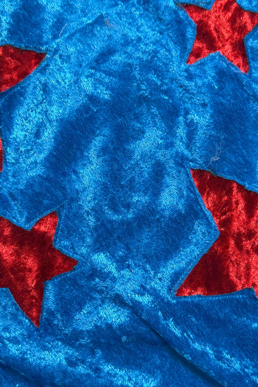 Close-up of a shiny Velvet fabric with a bold pattern. The fabric is primarily bright blue with cut-out red stars scattered across the surface, creating a textured contrast between the two colors.
