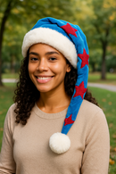 A young woman with medium brown skin and curly hair smiles warmly in a park, wearing a blue Velvet Santa-style hat with red stars, a white faux fur trim, and a fluffy white pom-pom.
