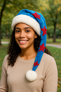 A young woman with medium brown skin and curly hair smiles warmly in a park, wearing a blue Velvet Santa-style hat with red stars, a white faux fur trim, and a fluffy white pom-pom.