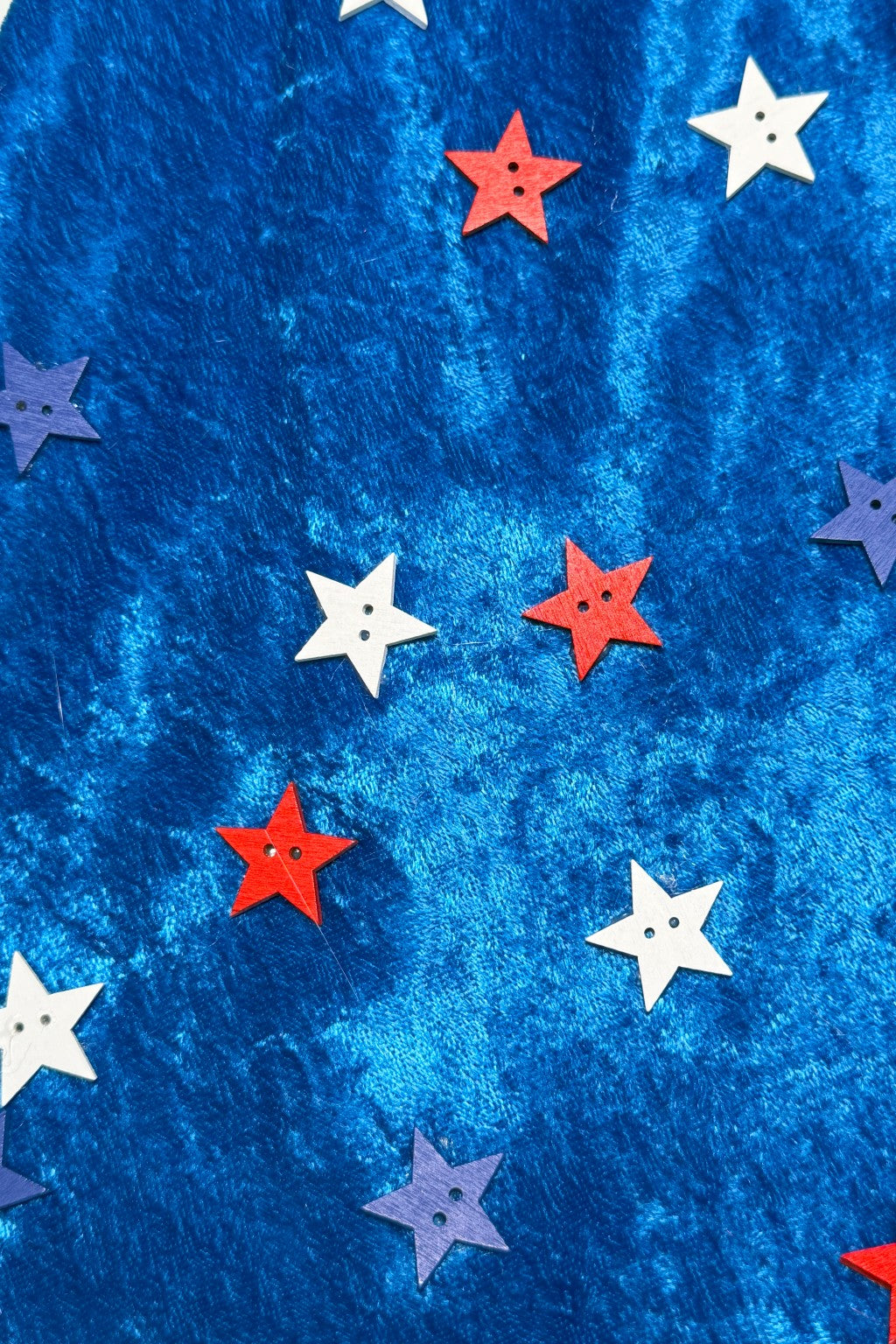 Pattern of red, white, and blue star-shaped buttons with two holes in the center, scattered across a shiny, textured royal blue fabric background.