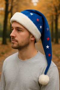 A young man with a short beard stands on an autumn path lined with golden trees, wearing a blue Santa-style hat with white faux fur trim, fluffy pom-pom, and red, white, and blue star decorations.