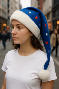 A young woman stands on a busy street wearing a white shirt and a festive blue Santa-style hat with white faux fur trim, fluffy pom-pom, and red, white, and blue star decorations.