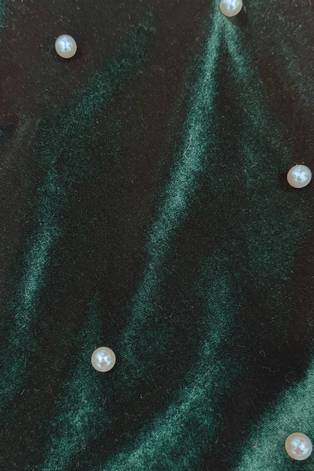 Close-up of dark green Velvet fabric with a wavy, iridescent sheen, decorated with small round pearl-like beads scattered across the surface.