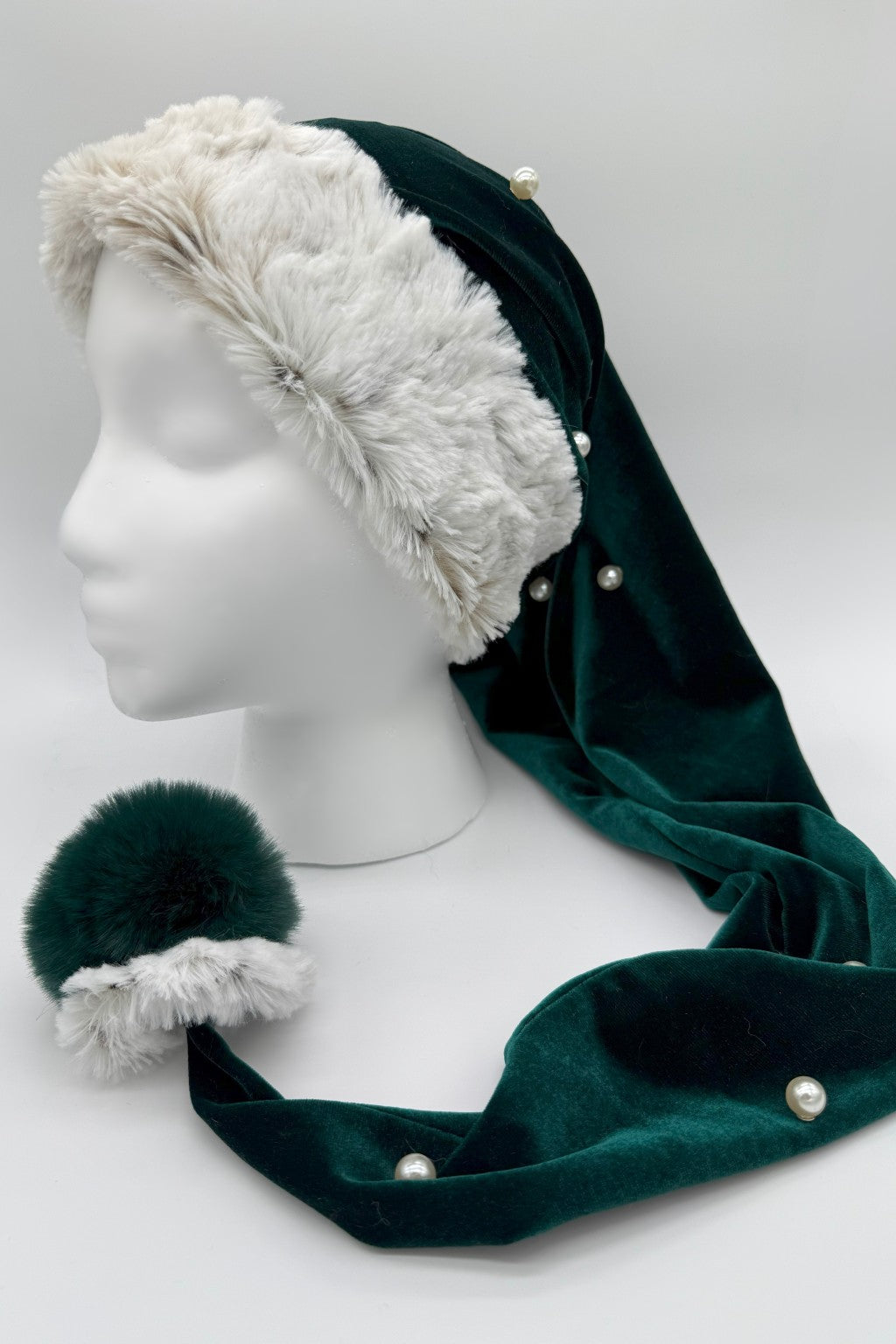 A dark green Velvet Santa-style hat with white faux fur trim and pearl accents, displayed on a white mannequin head. The hat has a long tail ending in a matching dark green and white faux fur pom-pom, shown resting on the surface.