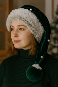 A young woman indoors wearing a deep green Velvet Santa-style hat with white faux fur trim, pearl accents along the tail, and a matching green-and-white faux fur pom-pom. She has light skin, short brown hair, and a calm expression, dressed in a dark green ribbed turtleneck.