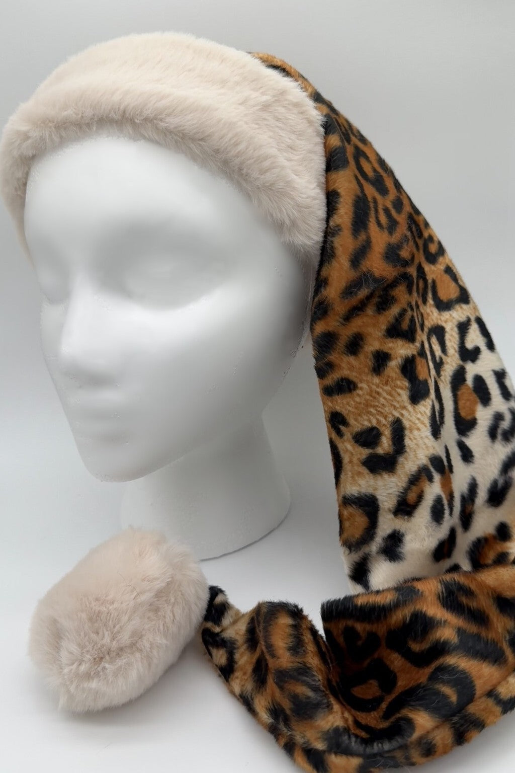 A faux fur leopard print hat with a long tail ending in a beige pom-pom, displayed on a white mannequin head. The hat has a wide beige fur trim around the forehead.